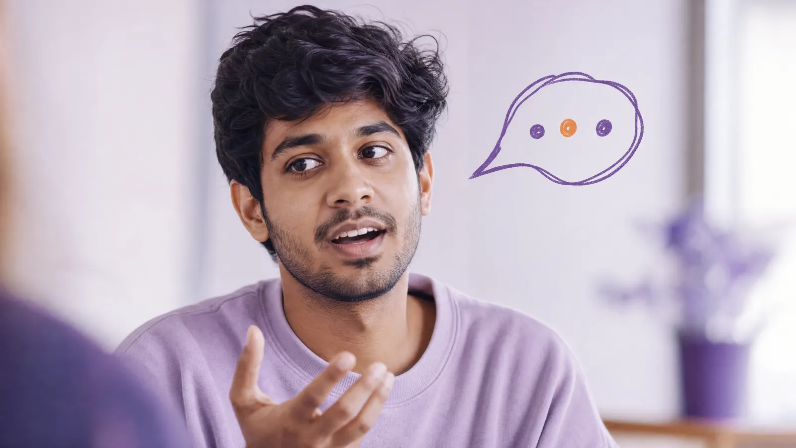 A young man pauses mid-sentence with his hand raised, searching for a word, a doodled speech bubble beside him holding three dots with the middle one orange